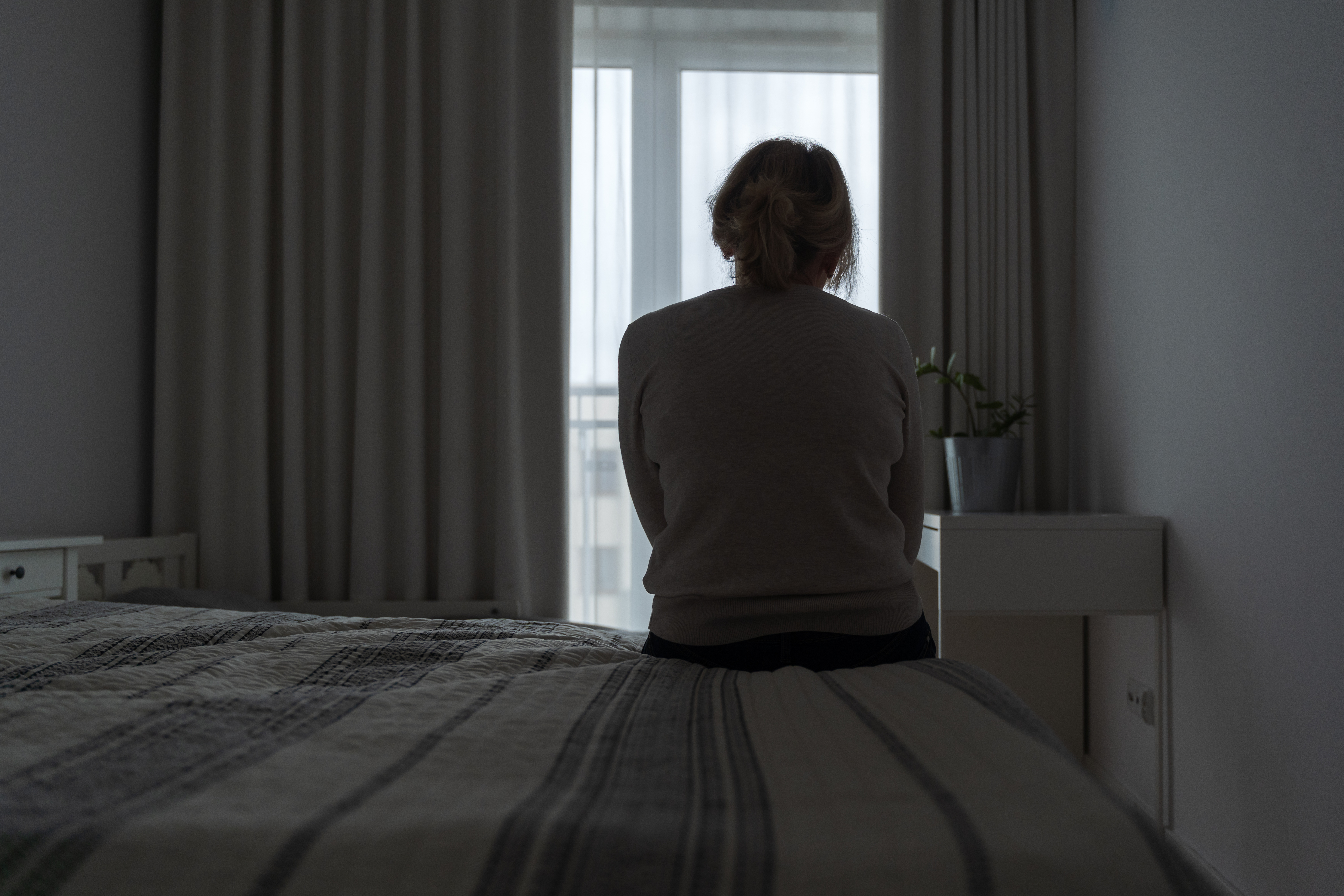 Frau sitzt mit dem Rücken zum Betrachter auf einer Bettkante und schaut nach vorne zum Fenster