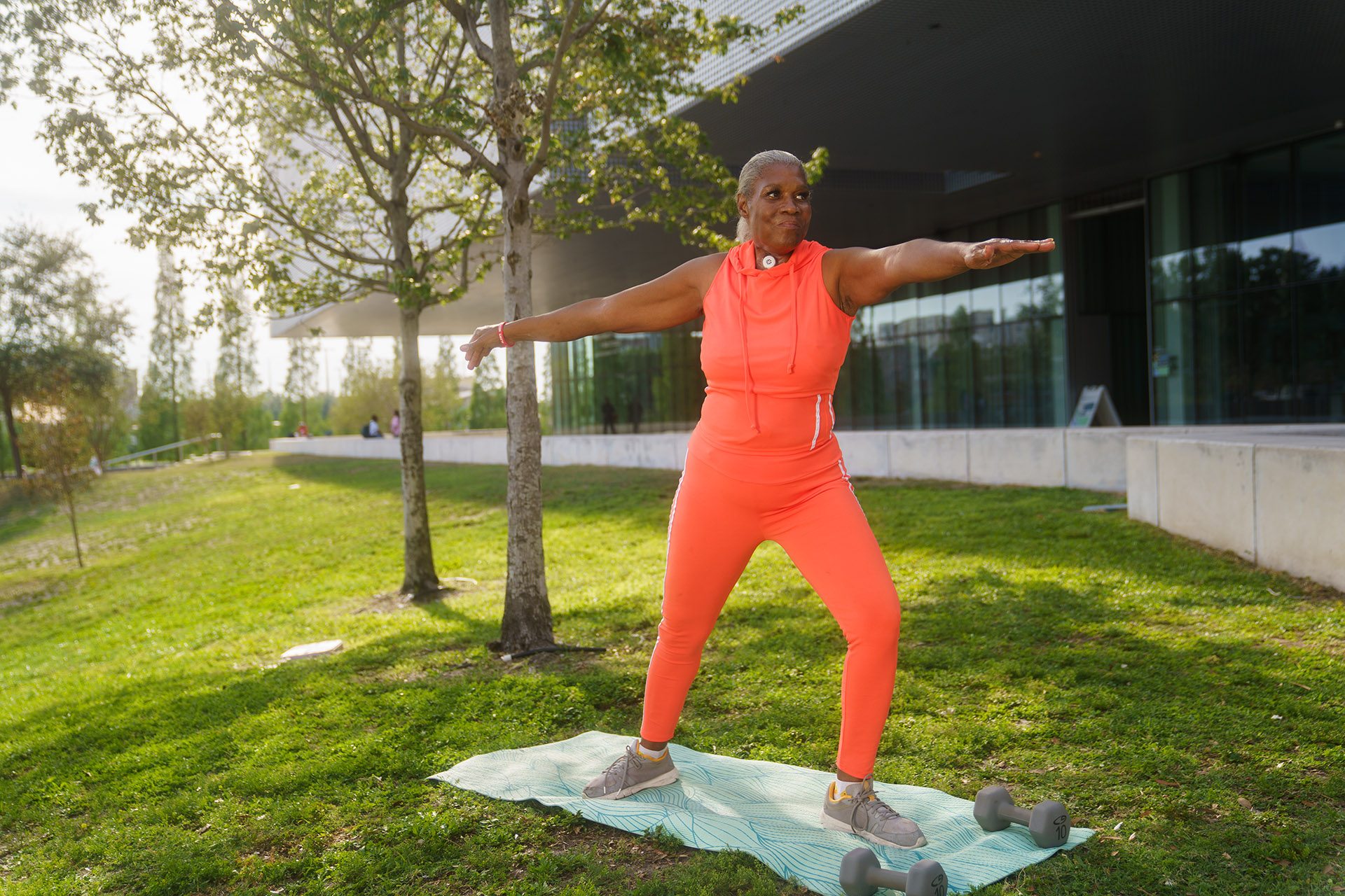 Laryngektomierte Frau steht auf einer Wiese auf einer Yogamatte und macht die Warrier Pose
